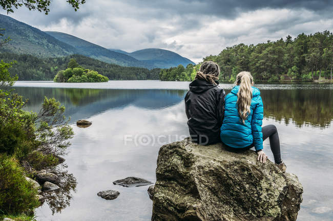 Paar sitzt auf Felsen und blickt auf ruhigen See, loch an eilein, Schottland — Stockfoto