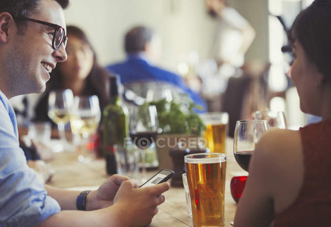 Pareja con teléfono celular hablando y bebiendo cerveza y vino en la mesa del restaurante - foto de stock