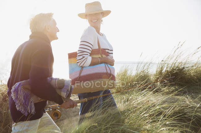 Sorrindo casal maduro com vara de pesca andando na grama ensolarada praia — Fotografia de Stock