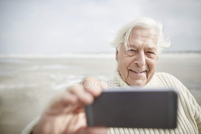 Hombre mayor sonriente tomando selfie con teléfono celular en la playa - foto de stock