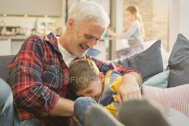 Juguetón padre cosquillas hijo en sala de estar sofá - foto de stock