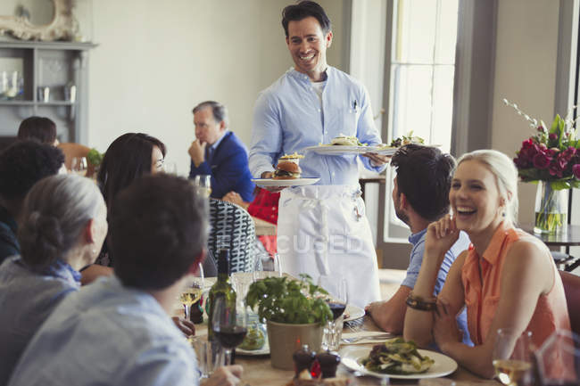 Усміхнений офіціант, який подає їжу друзям, обідаючи за столом ресторану — стокове фото