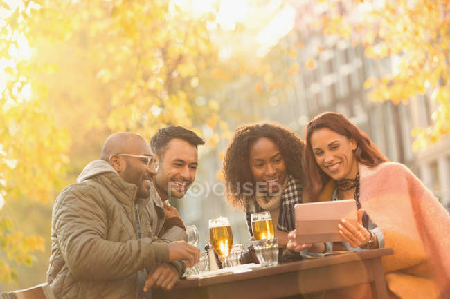 Улыбающиеся друзья пьют пиво и делают селфи с цифровым планшетом в осеннем кафе на тротуаре — стоковое фото