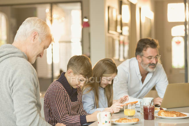 Male gay parents and children eating breakfast and using laptop and digital tablet at kitchen counter — Stock Photo