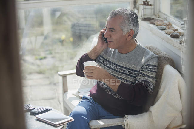 Senior man drinking coffee and talking on cell phone on sun porch — Stock Photo