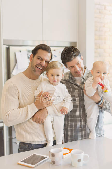 Portrait male gay parents holding baby sons in kitchen — Stock Photo