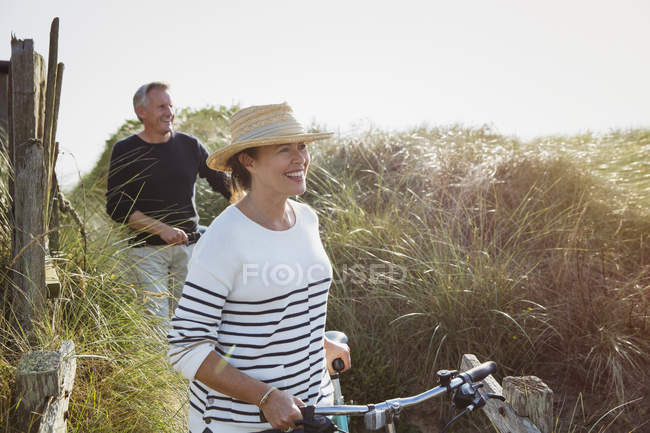 Mature couple walking bicycles along sunny beach grass — Stock Photo