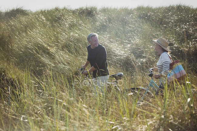 Mature couple walking bicycles in sunny beach grass — Stock Photo