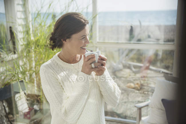 Усміхнена зріла жінка п'є каву на пляжному будиночку сонячний ганок — стокове фото
