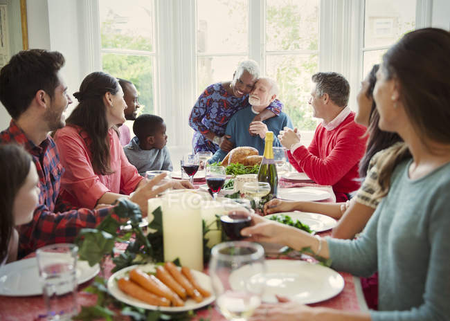 Cariñosa pareja de ancianos multiétnicos abrazándose en la mesa de la cena de Navidad familiar - foto de stock