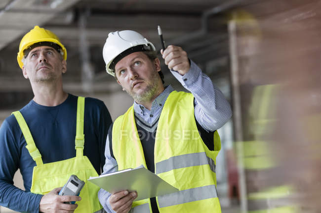 Hombre capataz e ingeniero con portapapeles mirando hacia arriba en el sitio de construcción - foto de stock
