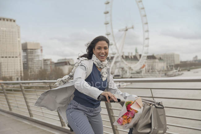 Portrait smiling woman bike riding on bridge over Thames River near Millennium Wheel, London, UK — Stock Photo