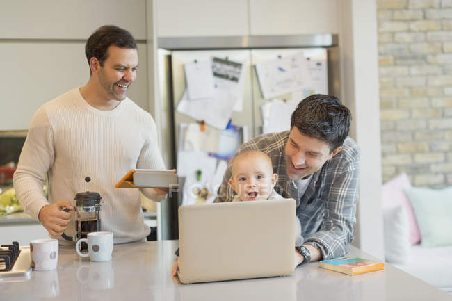 Male gay parents and baby son using laptop and digital tablet in kitchen — Stock Photo