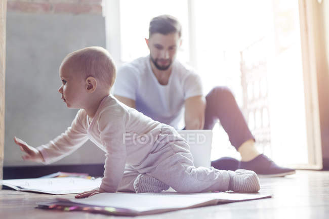 Малышка ползает по полу рядом с отцом, работающим за ноутбуком — стоковое фото