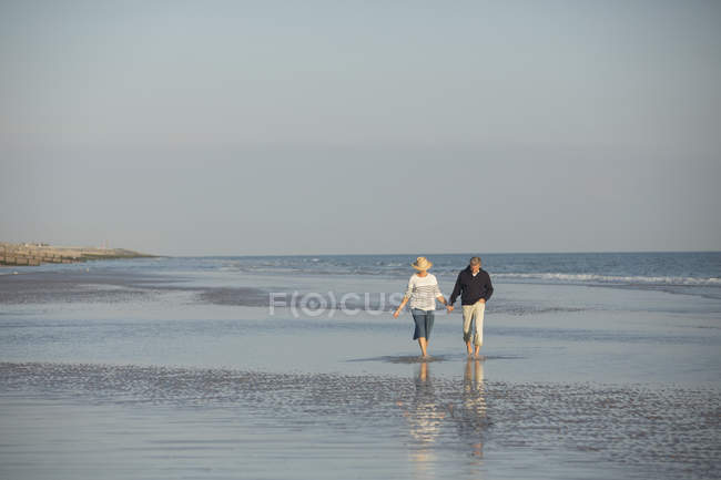 Pareja madura cogida de la mano caminando en el sol del océano playa surf - foto de stock