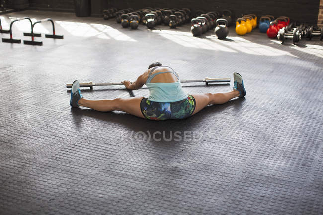 Junge Frau streckt sich mit gespreizten Beinen und benutzt Langhantel im Fitnessstudio — Stockfoto