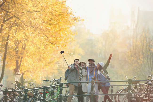 Jeunes amis ludiques prenant selfie avec bâton de selfie sur le pont d'automne urbain, Amsterdam — Photo de stock