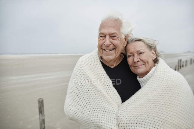 Sorridente affettuosa coppia anziana avvolta in una coperta sulla spiaggia — Foto stock