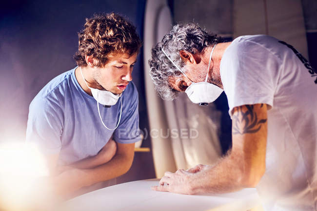 Focused men wearing protective masks sanding surfboard in workshop — Stock Photo