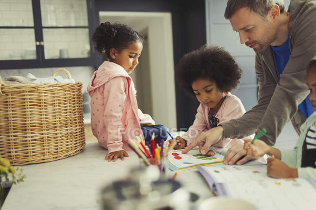 Padre e hijas multiétnicos para colorear y usar libros de actividades en la cocina - foto de stock