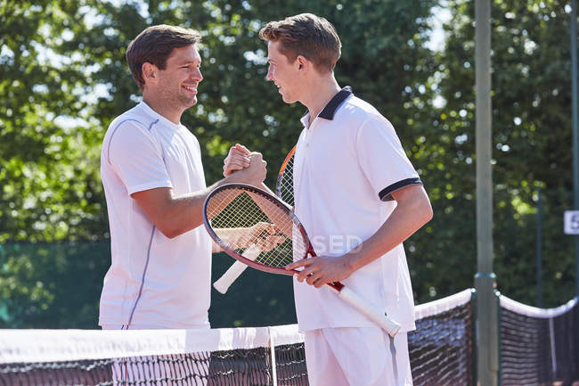 Sorrindo jovens jogadores de tênis do sexo masculino aperto de mão no sportsmanship sobre a rede na quadra de tênis — Fotografia de Stock