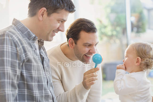 Male gay parents and baby son playing with rattle — Stock Photo