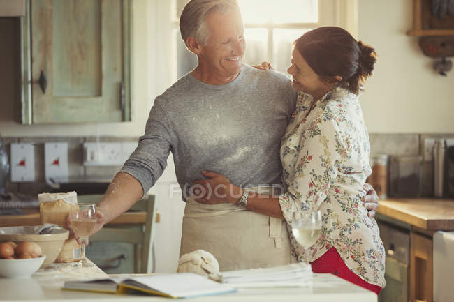 Affectueux couple d'âge mûr câlins, cuisson et boire du vin dans la cuisine — Photo de stock