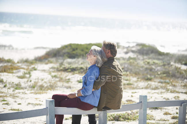 Affectionate senior couple sitting on sunny beach fence — Stock Photo