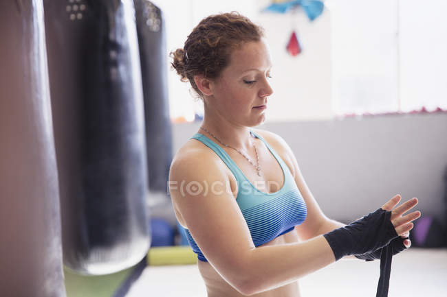 Boxerin wickelt Handgelenke neben Boxsack in Turnhalle — Stockfoto