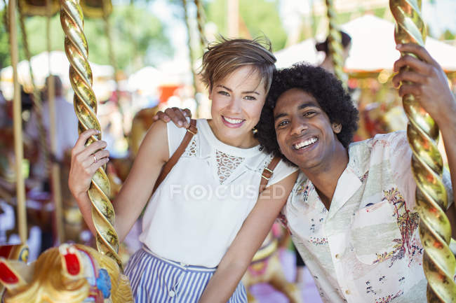Joven pareja multirracial sonriendo en el carrusel en el parque de atracciones - foto de stock