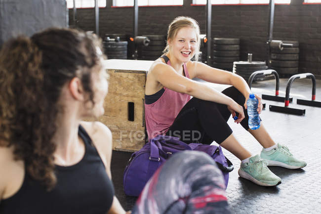 Улыбающиеся молодые женщины отдыхают и пьют воду после тренировки в тренажерном зале — стоковое фото