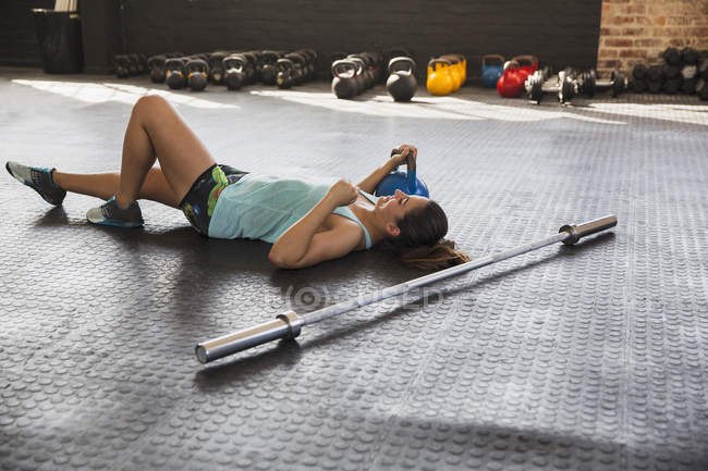 Молода жінка відпочиває, лежачи на підлозі спортзалу поруч з барбелом і чайником — стокове фото