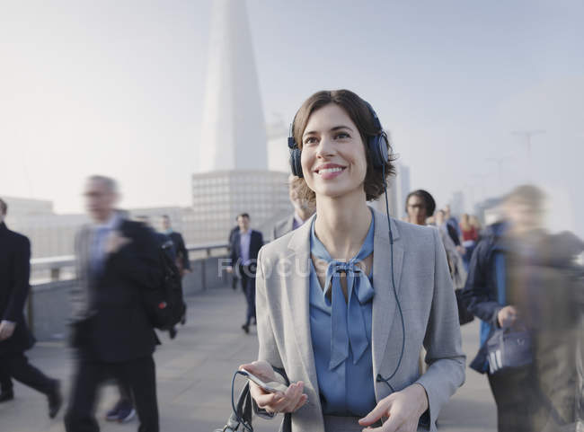 Mujer de negocios sonriente y confiada escuchando música con teléfono inteligente y auriculares en el puente peatonal urbano - foto de stock