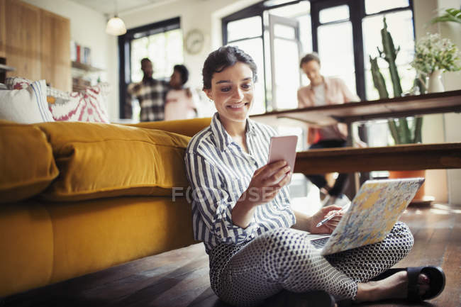 Mujer joven sonriente utilizando el ordenador portátil y mensajes de texto con teléfono inteligente en el piso de la sala de estar - foto de stock