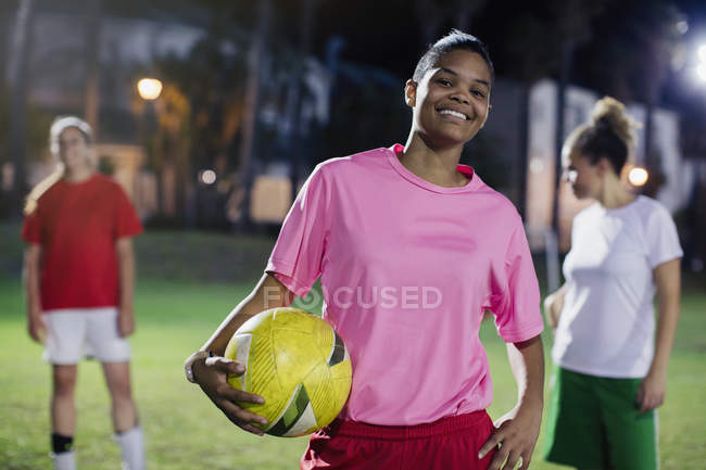 Портрет впевнена, усміхнена молода жінка-футболістка з м'ячем на полі вночі — стокове фото