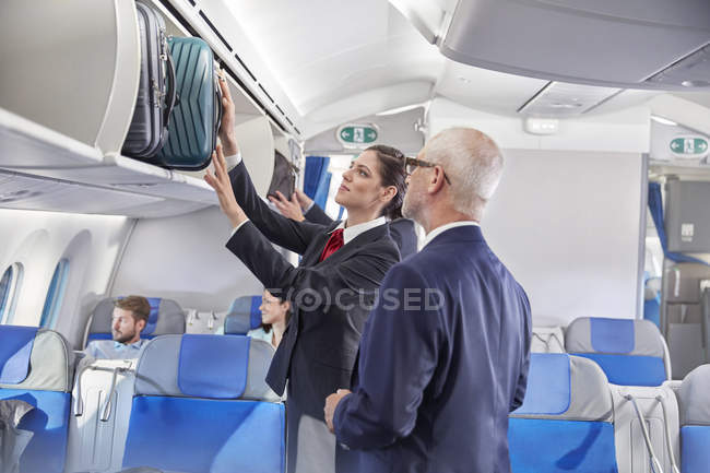 Assistente de bordo ajudando homem de negócios a colocar bagagem no compartimento aéreo no avião — Fotografia de Stock