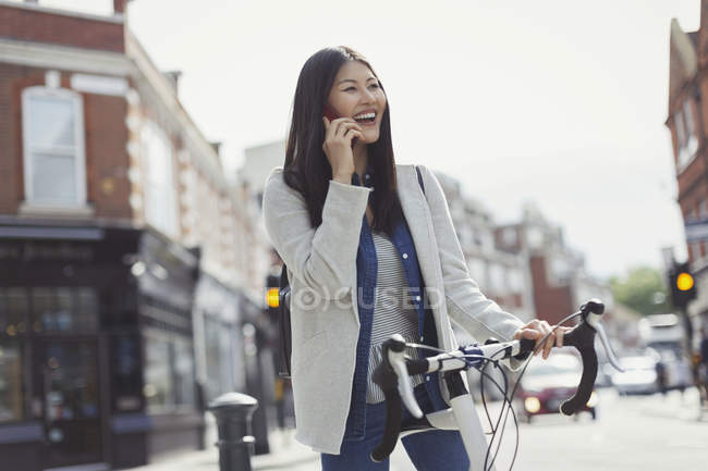 Une jeune femme souriante qui fait la navette à vélo, qui parle sur un téléphone portable dans une rue urbaine ensoleillée — Photo de stock