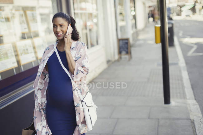Smiling pregnant woman talking on cell phone, walking along urban storefront — Stock Photo