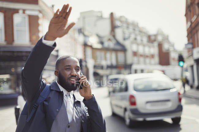 Young businessman talking on cell phone and hailing taxi on sunny urban street — Stock Photo