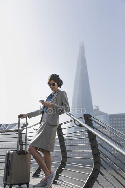 Businesswoman with suitcase listening to music with smart phone and headphones, London, UK — Stock Photo