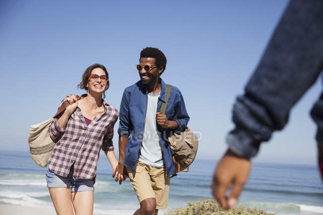 Улыбающаяся многонациональная пара, держащаяся за руки и идущая по солнечному летнему океанскому пляжу — стоковое фото