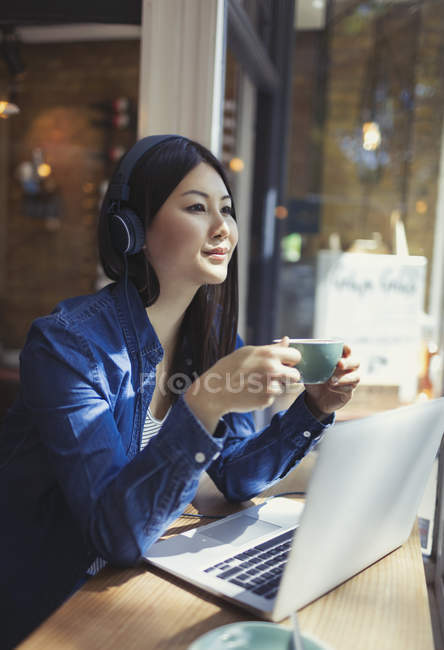 Pensive young woman listening to music with headphones and drinking coffee at laptop in cafe window — Stock Photo