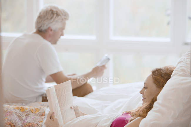 Libro de lectura de pareja y tableta digital en la cama - foto de stock