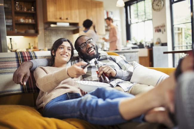Jeune couple souriant regardant la télévision, buvant de la bière et mangeant du pop-corn sur le canapé du salon — Photo de stock