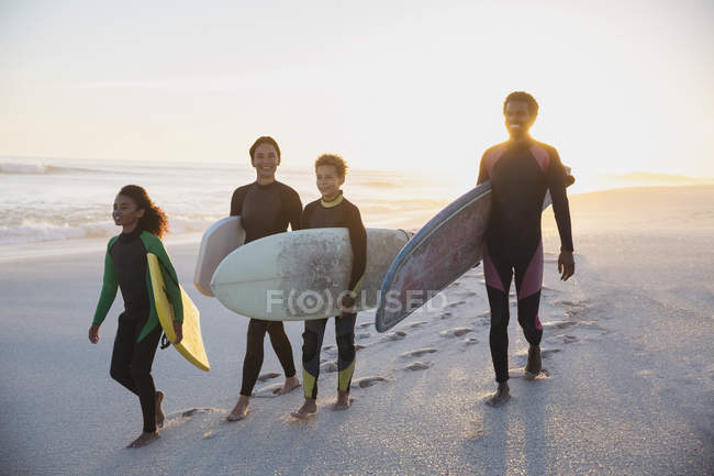 Surfistas familiares llevando tablas de surf en la playa del atardecer de verano - foto de stock