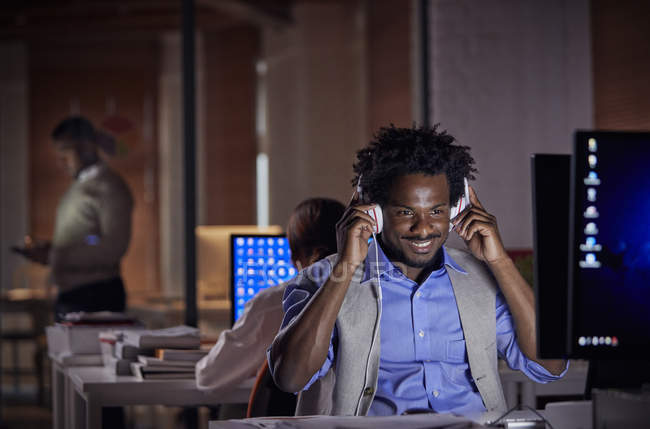 Hombre de negocios sonriente con auriculares escuchando música, trabajando hasta tarde en la computadora en la oficina oscura por la noche - foto de stock