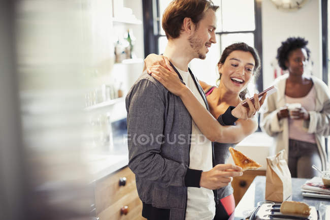 Affectionate couple hugging, texting with smart phone in kitchen — Stock Photo