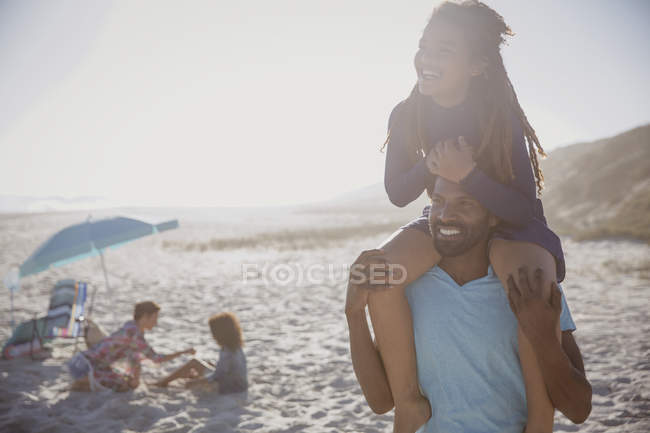 Улыбающийся отец несет дочь на плечах на солнечном летнем пляже — стоковое фото