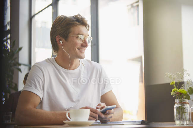 Hombre sonriente con auriculares y reproductor de mp3 escuchando música bebiendo café en la cafetería - foto de stock