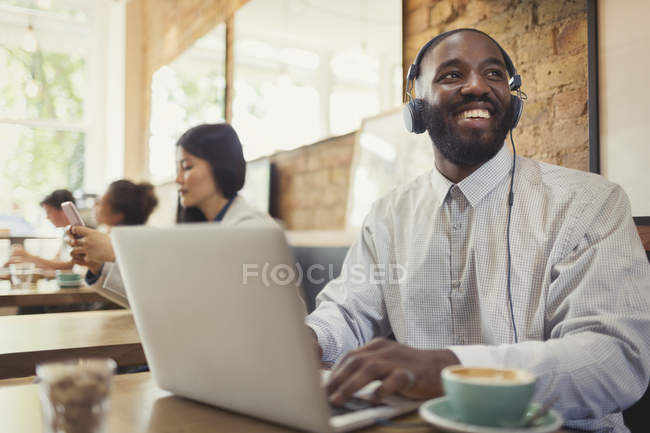 Un jeune homme souriant avec des écouteurs utilisant un ordinateur portable et buvant du café à la table du café — Photo de stock